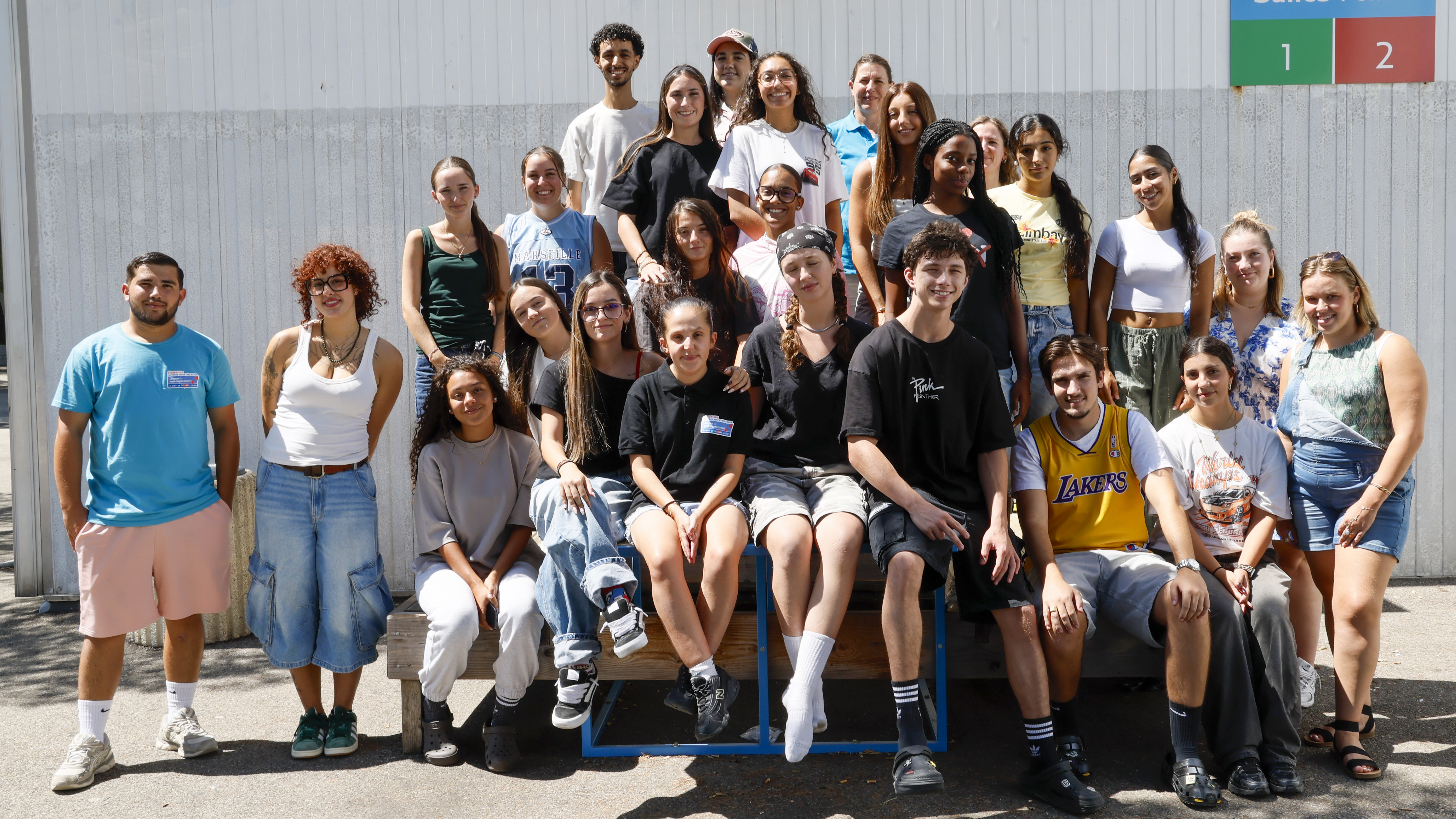 Il s'agit d'une photo de groupe en extérieur, prise en journée sous un soleil lumineux. Le groupe est composé d'une vingtaine de jeunes adultes (hommes et femmes d'une vingtaine d'années), disposés en plusieurs rangs devant un bâtiment. La plupart sourit à l'objectif dans une ambiance décontractée et estivale.