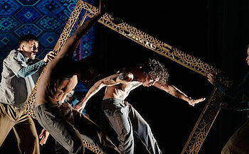 L’image montre quatre personnes en pleine performance sur scène. Trois d’entre elles sont torse nu ou partiellement vêtues, et portent des pantalons amples. Elles manipulent un grand cadre rectangulaire décoré de motifs ajourés dorés. Une personne au centre, penchée vers l’avant, semble en mouvement, avec les bras tendus et les muscles contractés. Deux autres personnes tiennent le cadre de chaque côté. En arrière-plan, on distingue un motif bleu et violet de style géométrique, probablement projeté ou faisant partie du décor. L’éclairage met en relief les corps et le cadre, créant une atmosphère dynamique et scénique.