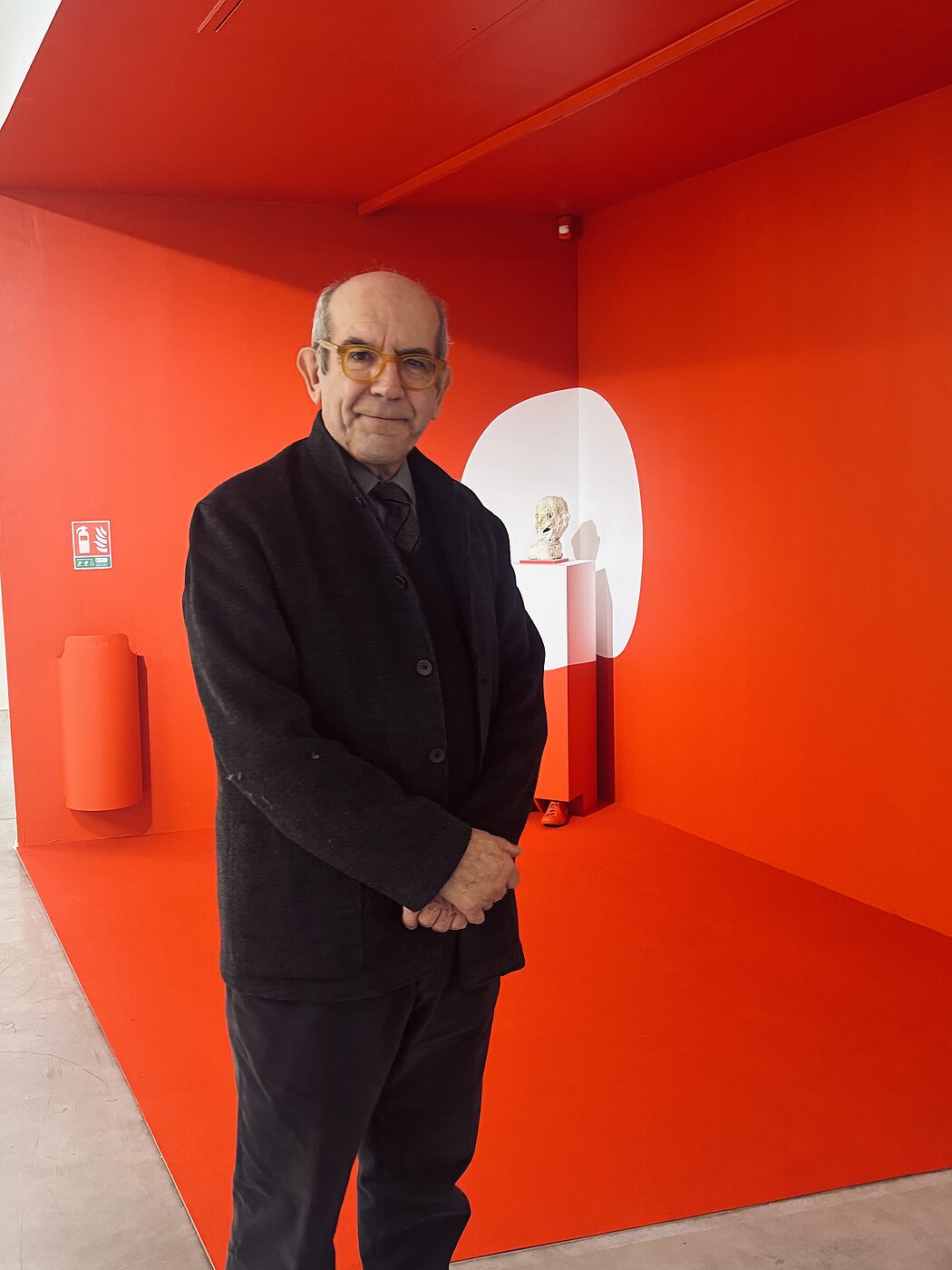 Cette image présente l'artiste italien **Gianni Dessì** posant au sein d'une installation artistique immersive et monochrome. L'œuvre joue sur des contrastes saisissants et une scénographie épurée.  Voici une description détaillée de la composition :  ### Le Sujet Principal  * **Gianni Dessì** se tient au premier plan, légèrement décentré vers la gauche. * Il arbore une expression sereine et un léger sourire. * Il est vêtu d'une **veste sombre texturée** boutonnée, sur un haut noir et une cravate à motifs discrets. Ses lunettes à monture jaune ambré apportent une touche de couleur chaude qui fait écho à l'arrière-plan.  ---  ### L'Environnement et l'Installation  * **Dominante Rouge :** Le décor est presque entièrement d'un **rouge vif et saturé**, couvrant le sol, les murs et même le plafond de l'alcôve. Cette uniformité crée une perte de repères spatiaux typique de certaines installations d'art contemporain. * **L'Élément Central :** En arrière-plan, une forme circulaire blanche semble avoir été "découpée" ou peinte sur l'angle du mur. À l'intérieur de cette zone blanche, une **petite sculpture de tête** (ressemblant à une forme organique ou géologique) repose sur un socle. * **Détails Insolites :** * On remarque un **pied chaussé d'une basket rouge** qui dépasse mystérieusement du bas du socle blanc, suggérant une présence cachée ou une extension de la sculpture. * À gauche, un extincteur et son boîtier ont été entièrement **peints en rouge** pour se fondre parfaitement dans la paroi, transformant un objet utilitaire en élément plastique.    ---  ### Analyse Visuelle  L'image capture un dialogue entre l'artiste et son œuvre. Le noir de sa tenue le détache nettement de l'arrière-plan flamboyant, tandis que les lignes de l'architecture et les formes géométriques (le cercle blanc, les angles droits) structurent l'espace autour de lui.