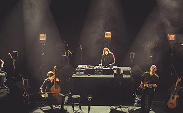 Photo des musiciens Bruno Allary, chant, guitares, Nolwenn Le Guern, viole de gambe, guitare basse, Clémence Niclas, chant, flûtes à bec et Mélissa Zantman, chant, percussions - L’image montre une scène faiblement éclairée sur laquelle se trouvent plusieurs musiciens. Au centre, une personne est debout derrière une table avec du matériel électronique, comme une table de mixage ou des platines. À gauche, une personne est assise et joue d’un violoncelle. À droite, une autre personne est debout et joue de la guitare électrique. Une guitare acoustique repose sur un support au sol. Plusieurs micros et projecteurs sont disposés autour de la scène. L’ambiance est sombre, avec des faisceaux de lumière éclairant les musiciens de manière ciblée 