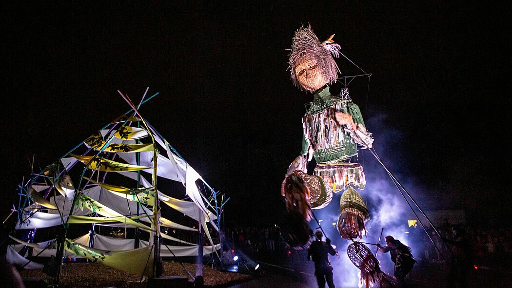Photo du spectacle la cabane à plume L’image montre une scène nocturne en extérieur.  À droite, une grande marionnette humanoïde est manipulée par plusieurs personnes visibles autour d’elle. Elle est composée de matériaux tressés et colorés, notamment du vert et du beige. Elle porte un oiseau sur la tête. La marionnette est tenue par des tiges longues, dirigées par les manipulateurs.  À gauche, une structure en forme de cabane ou tente est faite de bâches blanches et jaunes tendues entre des tiges ou poteaux colorés. La structure est éclairée par des lumières de différentes couleurs.  De la fumée ou du brouillard est visible au sol, surtout autour de la marionnette. En arrière-plan, on distingue une foule de personnes dans l'obscurité. Le ciel est noir, sans détail visible.