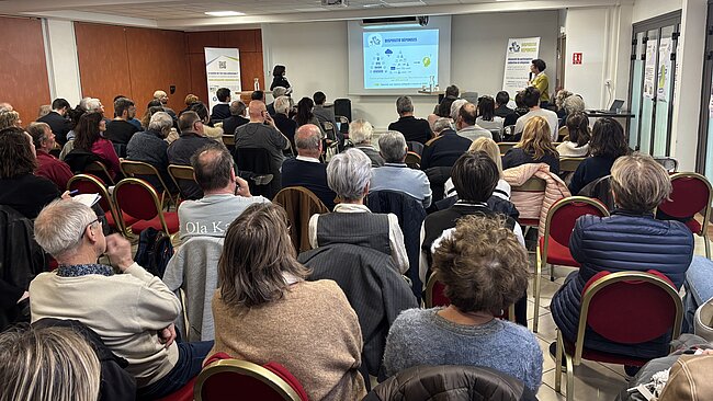 Cette photographie montre une réunion publique d'information se déroulant dans une salle de conférence ou une salle polyvalente. L'ambiance semble sérieuse et studieuse.  Voici une description détaillée de l'image : L'assistance      Composition : Une cinquantaine de personnes sont assises, de dos ou de profil par rapport à l'objectif. L'assemblée est majoritairement composée d'adultes et de seniors.      Disposition : Les participants sont installés sur des chaises rouges à structure dorée, disposées en rangées serrées faisant face à un écran de projection.  La présentation      Le contenu : Au fond de la salle, un écran projette une diapositive intitulée "DISPOSITIF RÉPONSES". On y voit des schémas, des logos de partenaires et des textes explicatifs sur la pollution de l'air et la participation citoyenne.      Les intervenants : À droite de l'écran, une personne debout avec un micro semble animer la séance. Une autre personne se tient à gauche de l'écran, près d'un pupitre.      Supports de communication : Deux "kakémonos" (enrouleurs publicitaires) encadrent la zone de présentation. Celui de droite porte le titre "Dispositif RÉPONSES" et explique la démarche de concertation autour de l'étang de Berre.