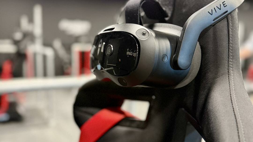 SALLE RV Casque de réalité virtuelle noir de marque "VIVE" accroché au dossier d'un siège baquet noir avec une ceinture rouge.  L'arrière-plan est flou, montrant des éléments métalliques et des lumières au plafond.