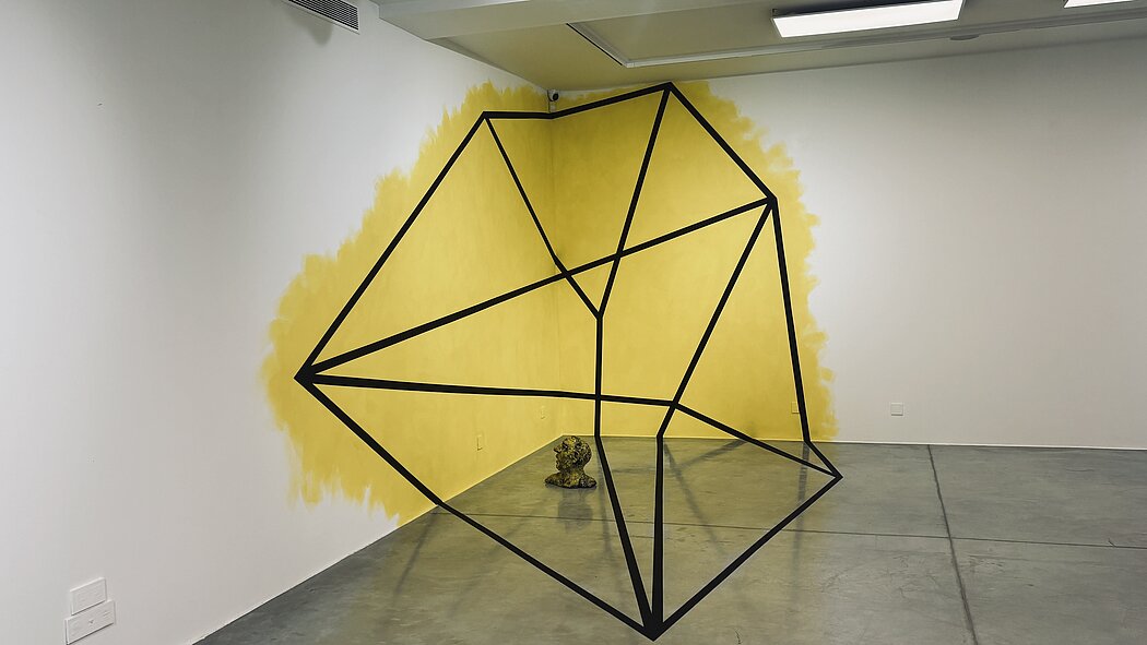 Cette image montre une installation artistique contemporaine située dans le coin d'une galerie aux murs blancs et au sol en béton lissé. L'œuvre joue sur la perspective, la géométrie et le contraste des couleurs.  Voici une description détaillée de la scène : Les Éléments Visuels      La forme géométrique : Une structure filiforme noire, ressemblant à un polyèdre ou à une cage de verre éclatée, semble "flotter" dans l'espace. En réalité, il s'agit d'une anamorphose : les lignes noires sont peintes directement sur les murs et le sol, mais elles ne s'alignent pour former cette figure parfaite que depuis un point de vue précis (celui du photographe).      L'arrière-plan jaune : Une large tache de peinture jaune vif, aux contours volontairement irréguliers et "brossés", occupe l'angle des deux murs. Elle sert de fond à la structure géométrique, la faisant ressortir avec force.      La sculpture au sol : Au centre de l'angle, sur le sol, repose une petite tête sculptée de couleur verdâtre/terreuse. Elle paraît minuscule et isolée à l'intérieur de la grande structure géométrique virtuelle.  Ambiance et Style      Minimalisme : La pièce est très épurée, typique d'un "cube blanc" de galerie d'art, ce qui concentre toute l'attention sur l'installation.      Jeu de perspective : L'œuvre défie la perception du spectateur en transformant des surfaces planes (murs et sol) en un objet qui semble posséder un volume tridimensionnel.