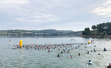 L'image montre un groupe de nageurs participant à une compétition ou un événement aquatique dans un plan d'eau. Les participants, qui portent des bonnets de bain de différentes couleurs, sont répartis sur la surface de l'eau. Certains sont en train de nager, tandis que d'autres semblent marcher ou se tenir debout dans l'eau. Un dispositif flottant de couleur jaune, probablement un repère de course, est visible au centre de l'image. En arrière-plan, on aperçoit des collines boisées et quelques personnes sur des kayaks ou des planches, surveillant l'événement. Le ciel est partiellement nuageux.