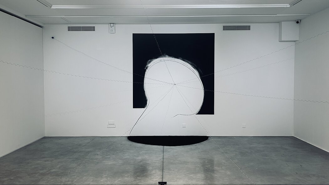 Cette œuvre de **Gianni Dessì**, intitulée ***Camera picta*** (réalisée vers 2008), est une installation immersive qui joue avec la perception spatiale et le dialogue entre la peinture et l'espace architectural.  Voici une description détaillée des éléments qui la composent :  ---  ### 1. La Figure Centrale : Un Portrait en Négatif  Au mur du fond, on observe une forme imposante qui suggère un **profil humain** ou une tête stylisée.  * **Contraste chromatique :** L'artiste utilise un contraste radical entre le noir et le blanc. La silhouette est "creusée" dans un carré noir, créant un effet de vide ou d'absence. * **Le traitement pictural :** Les bords ne sont pas nets ; on aperçoit des traces de pinceau blanches qui semblent déborder sur le noir, donnant une impression de vibration ou d'énergie autour du crâne. * **L'ombre au sol :** Au pied du mur, une demi-lune noire est peinte sur le sol, prolongeant la figure murale dans l'espace physique du spectateur et ancrant la silhouette dans la pièce.  ### 2. Le Réseau de Fils : L'Expansion de la Peinture  L'aspect le plus frappant est le réseau de **fils métalliques ou cordes spiralées** qui partent d'un point central situé sur le visage (probablement à l'emplacement de l'œil).  * **Rayonnement :** Ces fils se déploient en étoile dans toutes les directions de la salle : vers le plafond, les murs latéraux et les coins au sol. * **Ancrage :** Sur le sol, on remarque de petits objets (sortes de poids ou de bobines) qui maintiennent ces fils en tension. * **Lien physique :** Ce dispositif transforme une image bidimensionnelle (la peinture au mur) en une sculpture tridimensionnelle. Le spectateur ne regarde plus seulement une œuvre, il "entre" dedans.  ### 3. Composition et Symbolique  * **L'Anamorphose :** Dessì est connu pour ses jeux de points de vue. Selon l'endroit où l'on se place dans la salle, la figure peut paraître déformée ou, au contraire, s'aligner parfaitement. * **La Vision et la Pensée :** Le fait que tous les fils convergent vers l'œil ou le centre de la tête évoque la transmission de la pensée, la vision qui se projette vers l'extérieur ou, inversement, la manière dont le monde extérieur est capté par l'esprit. * **L'Espace "Picta" (Peint) :** Le titre fait référence à la *Chambre des Époux* de Mantegna (*Camera degli Sposi*), suggérant que l'artiste ne peint pas seulement sur une toile, mais qu'il "peint" la pièce entière avec des lignes de force.  ---  ### Résumé de l'expérience  L'œuvre crée une tension entre le **vide** (le blanc de la silhouette) et la **matière** (les fils métalliques). Elle donne l'impression que la figure au mur est en train de "tisser" l'espace autour d'elle, piégeant le spectateur dans son champ de vision.