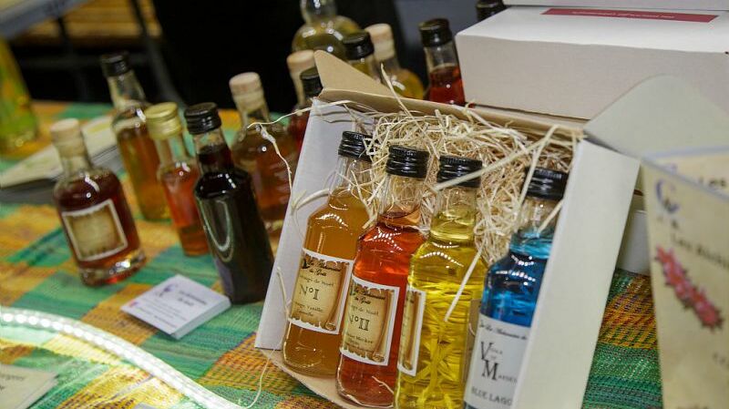 Cette image présente plusieurs bouteilles de liqueurs ou de boissons colorées. Les bouteilles sont de tailles variées et sont disposées sur un comptoir avec un fond multicolore. Certaines bouteilles sont rangées dans un carton, avec des étiquettes visibles comme « N°1 », « N°2 » et des variations de couleurs allant du jaune, orange, rouge au bleu. Le comptoir est décoré d'une nappe ou d’un tissu en textile, et des informations supplémentaires semblent être affichées à côté de certaines bouteilles.