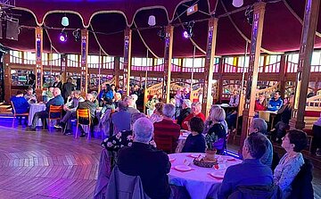 L'image montre un groupe de personnes assises à des tables rondes dans un espace intérieur. Les invités semblent être principalement des personnes âgées. L'environnement est un lieu décoré avec des murs en bois et des fenêtres colorées, créant une ambiance chaleureuse et conviviale. La lumière est tamisée, avec des spots de couleur violette qui ajoutent une touche festive. Certains invités regardent vers le devant, tandis que d'autres semblent discuter entre eux. On peut voir des décorations sur les tables, comme des plantes et des assiettes. Le lieu semble être un espace pour une réception ou un événement spécial.