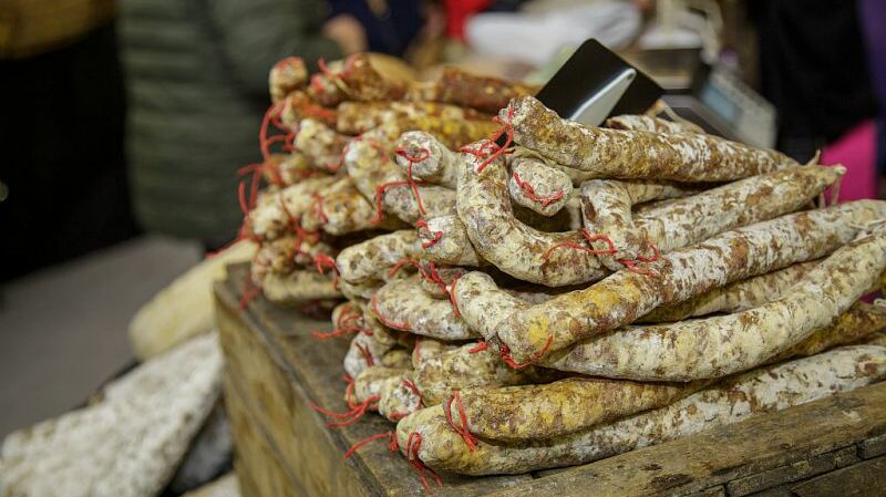 Cette image montre plusieurs saucissons suspendus et empilés sur un étal. Les saucissons sont recouverts d'une couche de moisissure blanche, typique des produits secs, et sont attachés avec des ficelles rouges. En arrière-plan, on aperçoit des gens, suggérant un marché ou un événement, et un téléphone portable est visible parmi les produits. L’étal semble être en bois, et l'ensemble de la scène évoque une ambiance artisanale et traditionnelle.