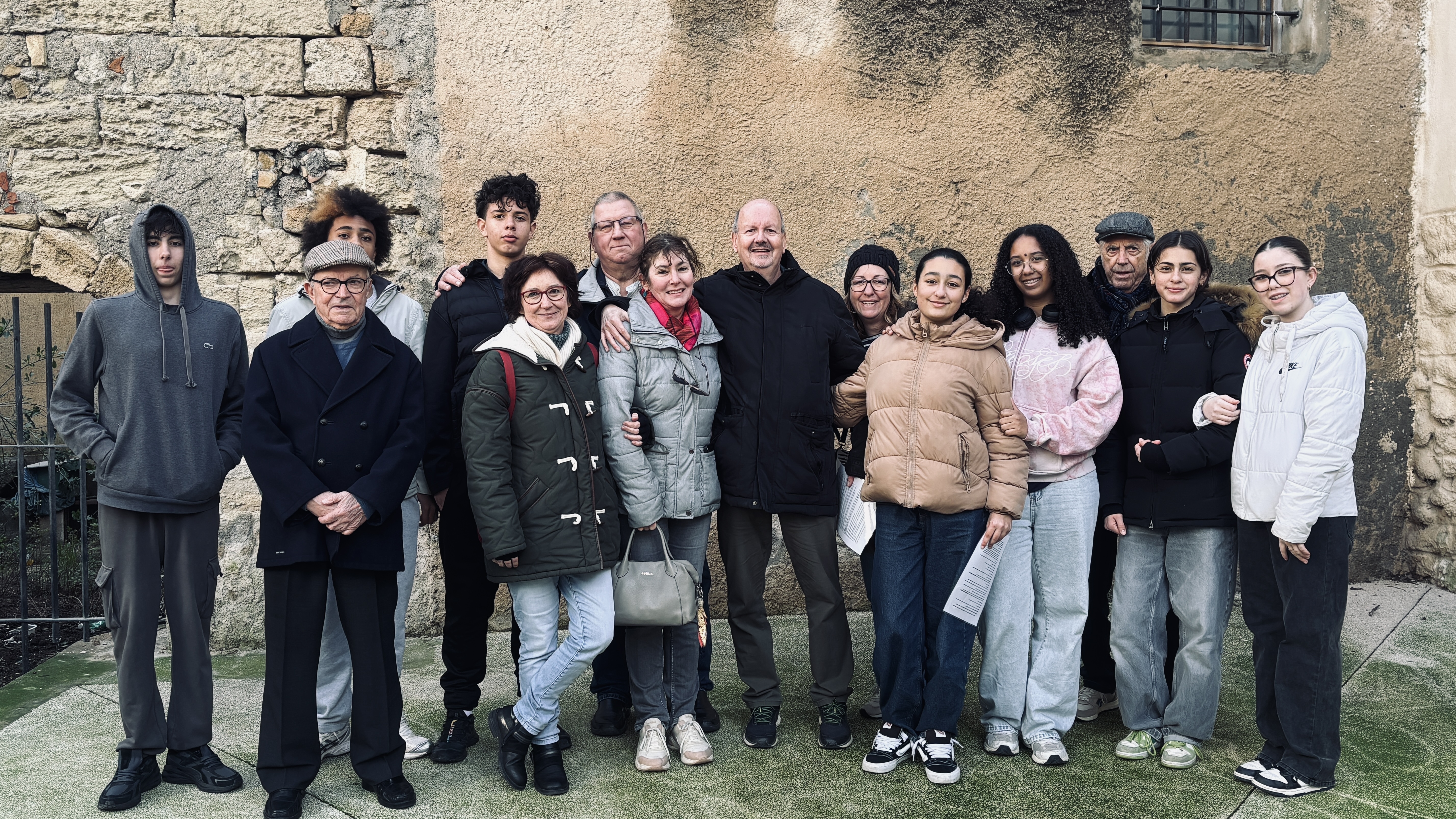     Requête réussie  Gemini said  Cette photo montre un groupe de treize personnes posant ensemble en extérieur, devant un mur de pierre ancienne et de crépi. L'ambiance semble être celle d'une réunion familiale ou amicale par un temps frais.  Voici une description plus détaillée de l'image : Le Groupe  Le groupe est composé d'hommes et de femmes de différents âges. Ils sont disposés sur deux rangs informels :      Au centre : Un homme avec un manteau noir semble être le point central, entouré de plusieurs femmes qui sourient.      À gauche : On remarque un homme plus âgé portant une casquette et un caban bleu marine, ainsi qu'un jeune homme en sweat à capuche gris.      À droite : Plusieurs jeunes femmes et un homme plus âgé avec une casquette plate complètent le groupe.      Interactions : Plusieurs personnes se tiennent par l'épaule ou le bras, ce qui suggère une grande proximité et une atmosphère chaleureuse.