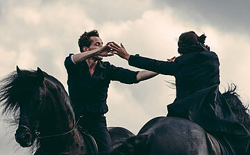 L'image montre deux personnes vêtues de noir, chacune montée sur un cheval noir. Elles sont engagées dans un mouvement chorégraphié ou expressif, les bras tendus l’un vers l’autre. Le ciel en arrière-plan est nuageux, avec une lumière diffuse. Les chevaux sont capturés en mouvement, et la scène transmet une impression de dynamisme et de tension.