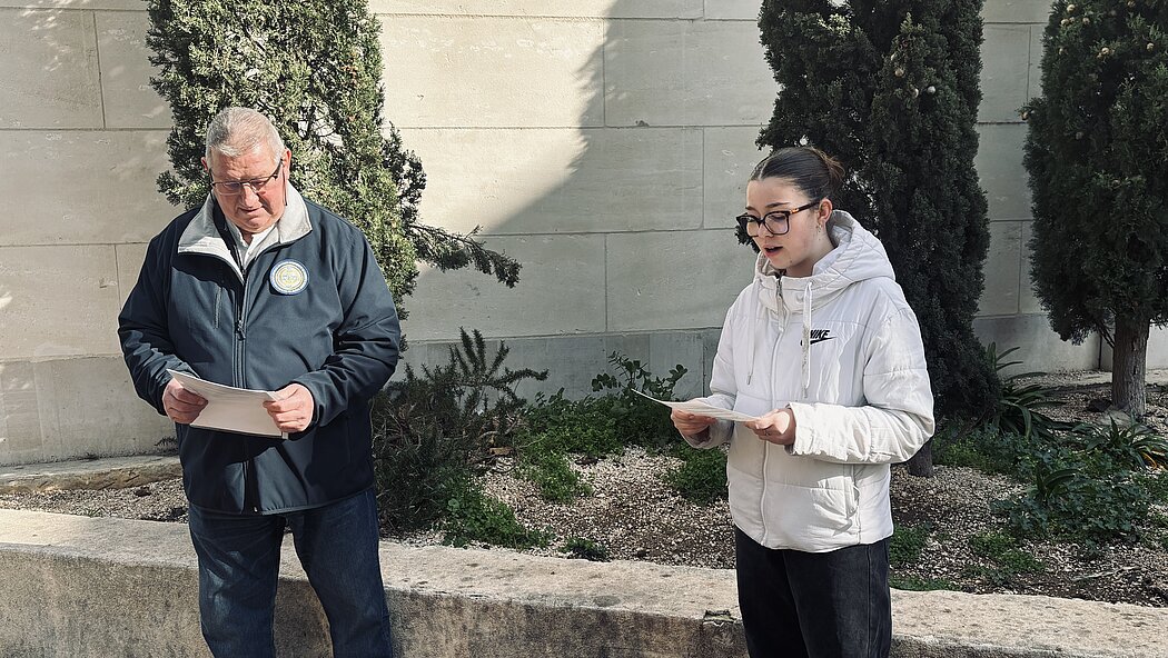 Cette image montre deux personnes se tenant côte à côte à l'extérieur, devant un mur en pierre claire. Les Personnes      À gauche : Un homme plus âgé, portant des lunettes, une veste bleu marine avec un écusson rond sur la poitrine, et un jean bleu. Il tient une feuille de papier et semble la lire attentivement.      À droite : Une jeune fille avec des lunettes et les cheveux attachés, vêtue d'une doudoune blanche à capuche de marque Nike et d'un pantalon noir. Elle tient également une feuille de papier et semble être en train de parler ou de lire à haute voix.  Le Décor      Arrière-plan : Un mur de blocs de pierre lisse et rectangulaire. Plusieurs arbres fins et sombres (ressemblant à des cyprès) sont plantés derrière eux.      Premier plan : Les deux personnes se tiennent derrière un muret en pierre basse qui délimite un petit espace de jardinage avec quelques plantes vertes et de la terre.  L'ambiance suggère un moment solennel ou une lecture publique...