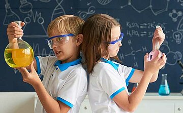 Sur l’image, on voit deux enfants vêtus de polos blancs avec des liserés bleus. Ils portent des lunettes de protection transparentes et bleues.  L’enfant à gauche tient un grand ballon de verre contenant un liquide jaune.  L’enfant à droite tient un ballon de verre plus petit contenant un liquide rose.  Ils se tiennent dos à dos.  En arrière-plan, il y a un tableau bleu foncé avec des dessins et formules scientifiques tracés à la craie blanche.  Sur une table derrière eux, on distingue d’autres récipients de laboratoire, dont un erlenmeyer contenant un liquide bleu.