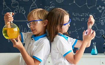 Sur l’image, on voit deux enfants vêtus de polos blancs avec des liserés bleus. Ils portent des lunettes de protection transparentes et bleues.  L’enfant à gauche tient un grand ballon de verre contenant un liquide jaune.  L’enfant à droite tient un ballon de verre plus petit contenant un liquide rose.  Ils se tiennent dos à dos.  En arrière-plan, il y a un tableau bleu foncé avec des dessins et formules scientifiques tracés à la craie blanche.  Sur une table derrière eux, on distingue d’autres récipients de laboratoire, dont un erlenmeyer contenant un liquide bleu.
