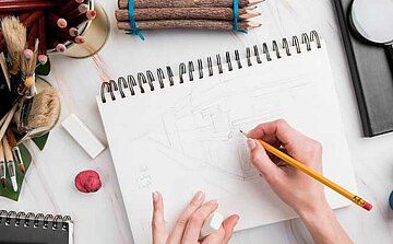 L'image montre un espace de travail créatif avec divers outils artistiques. Au centre, un carnet de croquis ouvert montre un dessin en cours, avec une main tenant un crayon jaune, en train de dessiner. À côté du carnet, on trouve une gomme blanche, ainsi que des blocs de papier et de petits pots de pinceaux et de crayons de couleur. Il y a aussi un bâton de modelage rouge et des bâtons de bois attachés par un élastique. En arrière-plan, des pinceaux sont regroupés, et plusieurs autres objets sont visibles, créant un environnement propice à la créativité et au travail artistique.