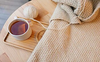 L'image montre une scène calme et cosy. Sur une table en bois clair, on voit une tasse de thé avec une teinte ambrée, placée sur un plateau en bois. À côté, il y a un rouleau de laine de couleur claire, et un morceau de tissu tricoté, probablement en laine également, qui semble être en train d'être travaillé. Le tout est posé sur une surface lisse, probablement en bois clair, avec des détails subtils du tricot visibles sur le tissu. L'ambiance dégagée est chaleureuse et confortable, suggérant un moment de détente et de créativité.