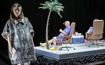 L’image montre trois personnes sur une scène. Au premier plan, une femme debout porte un vêtement ample de couleur grise et tient des feuilles de papier. À l’arrière-plan, deux personnes sont assises sur des chaises de bureau marron, sur une plateforme surélevée. Elles portent des vêtements violets et des chaussures jaunes. Une fausse palme éclairée est plantée au centre de la plateforme, à côté d’une glacière bleue et d’objets divers. Le sol autour de la plateforme est recouvert de nombreuses boules noires. L’arrière-plan est sombre.