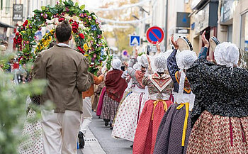 défilé de personnes en costumes traditionnels, marchant dans une rue. Un homme portant une veste beige porte une couronne florale, tandis que des femmes vêtues de robes anciennes et de bonnets blancs, typiques des costumes folkloriques, participent également à la procession. Les costumes sont colorés et très détaillés, et le décor montre une rue avec des panneaux de circulation et des bâtiments urbains en arrière-plan.