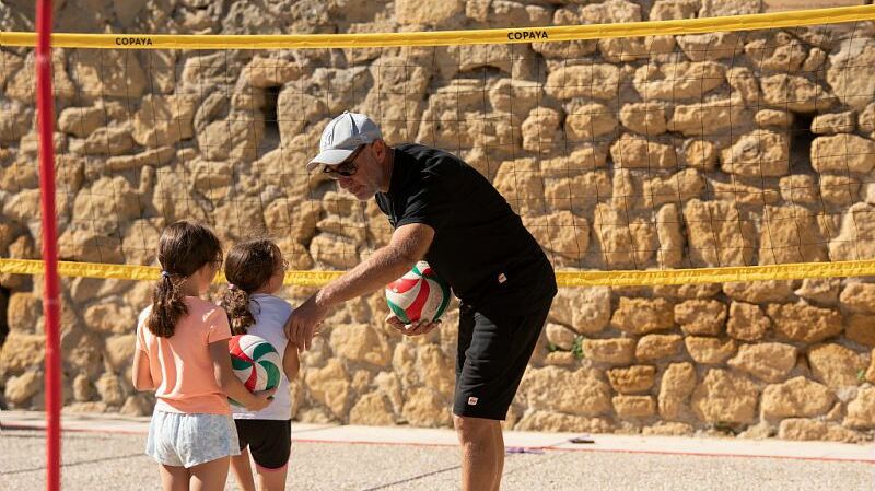 Un homme remet des ballons de volley à deux jeunes enfants sur un terrain de sport avec un filet, près d’un mur en pierre.