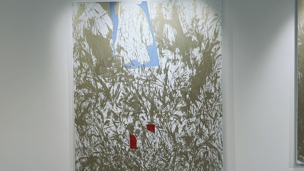 Cette œuvre est une peinture abstraite de grand format, accrochée sur un mur blanc de galerie. Elle se distingue par un mélange de textures brutes et de contrastes de couleurs primaires sur un fond neutre.  Voici une description détaillée de ses éléments :  ### Composition et Texture  * **Le Fond :** La majeure partie de la toile est recouverte d'un réseau dense et chaotique de coups de pinceau ou de traces de spatule de couleur **bronze/doré mat** sur un fond blanc cassé. Ces marques évoquent des herbes hautes, des ronces ou une écriture gestuelle très énergique. * **La Gestuelle :** L'application de la peinture semble instinctive et physique, créant une impression de mouvement organique qui remplit presque tout l'espace visuel.  ### Éléments Géométriques et Couleurs  Trois zones de couleurs unies viennent rompre cette texture broussailleuse :  1. **La Forme Bleue (Haut) :** Une forme bleu ciel, rappelant une fenêtre inversée ou un vêtement stylisé, occupe le centre supérieur. Elle semble "flotter" derrière ou à travers les marques dorées. 2. **Les Formes Rouges (Bas) :** Deux petites taches rouge vif, de formes irrégulières et angulaires, sont placées vers le bas au centre. Elles agissent comme des points d'ancrage visuel, attirant immédiatement l'œil par leur intensité.  ### Ambiance Générale  L'œuvre joue sur le contraste entre le **chaos des lignes dorées** et la **clarté des aplats de couleur**. Il s'en dégage une impression de paysage abstrait, où des éléments structurés (le bleu et le rouge) tentent d'émerger d'une nature sauvage ou d'un tumulte graphique.