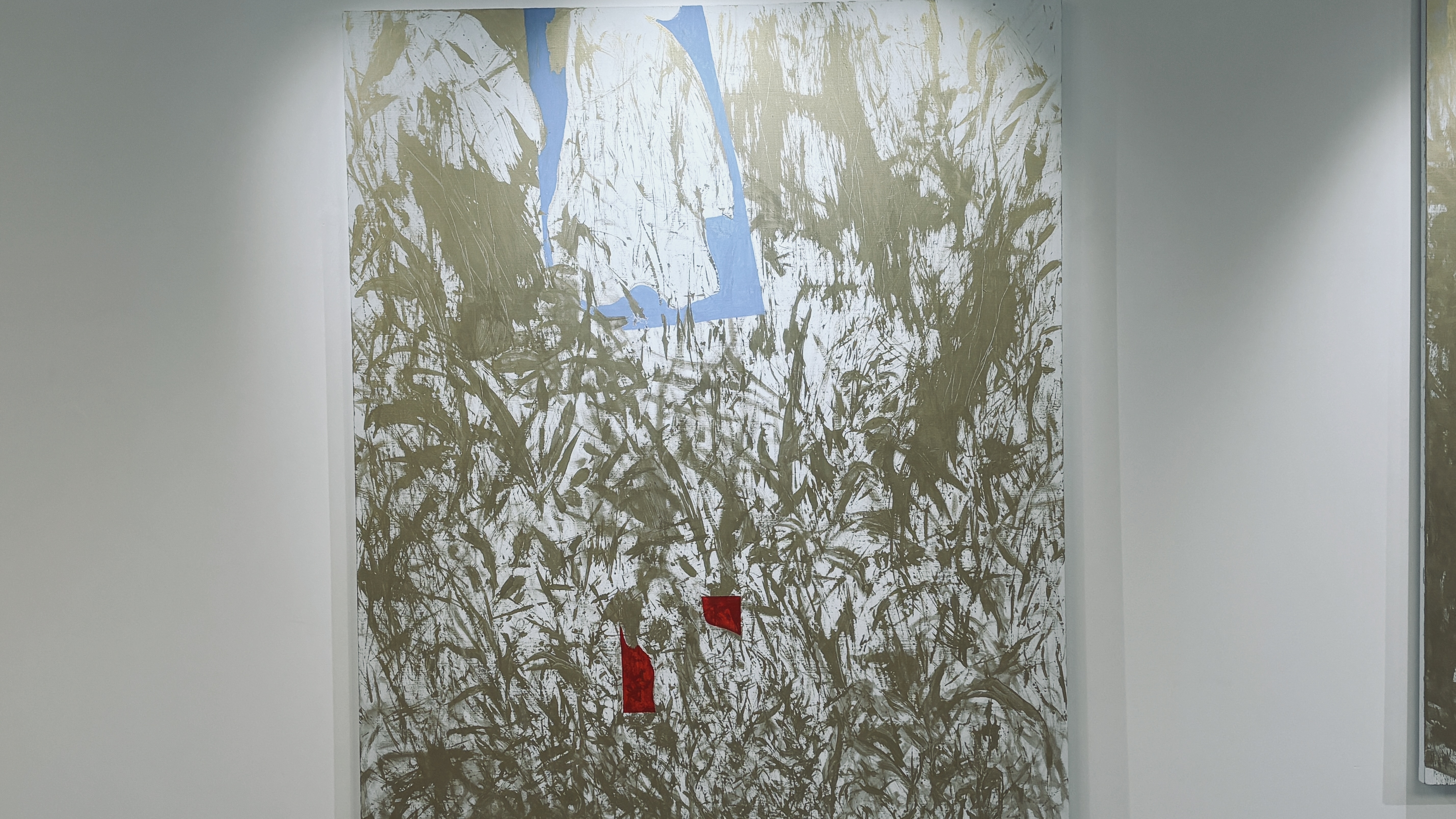 Cette œuvre est une peinture abstraite de grand format, accrochée sur un mur blanc de galerie. Elle se distingue par un mélange de textures brutes et de contrastes de couleurs primaires sur un fond neutre.  Voici une description détaillée de ses éléments :  ### Composition et Texture  * **Le Fond :** La majeure partie de la toile est recouverte d'un réseau dense et chaotique de coups de pinceau ou de traces de spatule de couleur **bronze/doré mat** sur un fond blanc cassé. Ces marques évoquent des herbes hautes, des ronces ou une écriture gestuelle très énergique. * **La Gestuelle :** L'application de la peinture semble instinctive et physique, créant une impression de mouvement organique qui remplit presque tout l'espace visuel.  ### Éléments Géométriques et Couleurs  Trois zones de couleurs unies viennent rompre cette texture broussailleuse :  1. **La Forme Bleue (Haut) :** Une forme bleu ciel, rappelant une fenêtre inversée ou un vêtement stylisé, occupe le centre supérieur. Elle semble "flotter" derrière ou à travers les marques dorées. 2. **Les Formes Rouges (Bas) :** Deux petites taches rouge vif, de formes irrégulières et angulaires, sont placées vers le bas au centre. Elles agissent comme des points d'ancrage visuel, attirant immédiatement l'œil par leur intensité.  ### Ambiance Générale  L'œuvre joue sur le contraste entre le **chaos des lignes dorées** et la **clarté des aplats de couleur**. Il s'en dégage une impression de paysage abstrait, où des éléments structurés (le bleu et le rouge) tentent d'émerger d'une nature sauvage ou d'un tumulte graphique.
