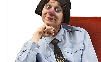 Sur cette image, on voit une personne maquillée en clown, assise sur un fauteuil rouge. Elle porte une chemise bleu clair boutonnée et une cravate noire ornée d’un motif représentant un clavier d’ordinateur avec la touche "ESC" en évidence. Son maquillage se compose d’un nez rouge arrondi, de grands cercles noirs autour des yeux et d’un rouge à lèvres débordant légèrement des lèvres, formant un sourire accentué.  Elle porte également un bonnet noir souple couvrant partiellement ses cheveux. Sa tête est légèrement inclinée, et son menton repose sur son poing fermé, dans une posture détendue, presque pensive. Le fond est blanc, mettant en valeur le personnage au centre de l’image.