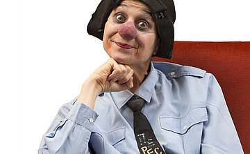 Sur cette image, on voit une personne maquillée en clown, assise sur un fauteuil rouge. Elle porte une chemise bleu clair boutonnée et une cravate noire ornée d’un motif représentant un clavier d’ordinateur avec la touche "ESC" en évidence. Son maquillage se compose d’un nez rouge arrondi, de grands cercles noirs autour des yeux et d’un rouge à lèvres débordant légèrement des lèvres, formant un sourire accentué.  Elle porte également un bonnet noir souple couvrant partiellement ses cheveux. Sa tête est légèrement inclinée, et son menton repose sur son poing fermé, dans une posture détendue, presque pensive. Le fond est blanc, mettant en valeur le personnage au centre de l’image.