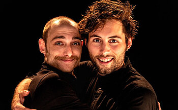 L’image montre deux personnes habillées en noir, en train de s’enlacer. Elles sont placées au centre de l’image, regardant toutes les deux vers l’objectif, avec un sourire. La personne à gauche a le crâne rasé sur le dessus avec un peu de cheveux sur les côtés, une moustache fine et des sourcils marqués. La personne à droite a des cheveux épais, bruns et en bataille, ainsi qu'une barbe courte. L’arrière-plan est noir, ce qui met en évidence les visages et les expressions des deux individus grâce à un éclairage chaud dirigé sur eux.