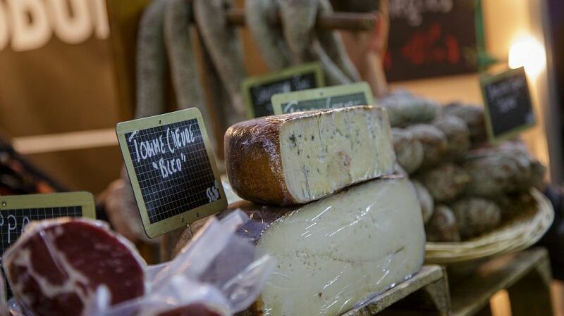 Cette image montre une rangée de fromages de différentes formes et tailles, présentée sur une table en bois. Les fromages sont bien organisés, certains sont coupés en tranches et d'autres restent intacts, chacun ayant des textures et des croûtes distinctes. Des étiquettes sont visibles, indiquant probablement les types de fromages. En arrière-plan, on peut apercevoir des petits accessoires comme des couteaux à fromage, et une ambiance de marché ou de foire est présente avec des clients qui semblent s'intéresser aux produits.