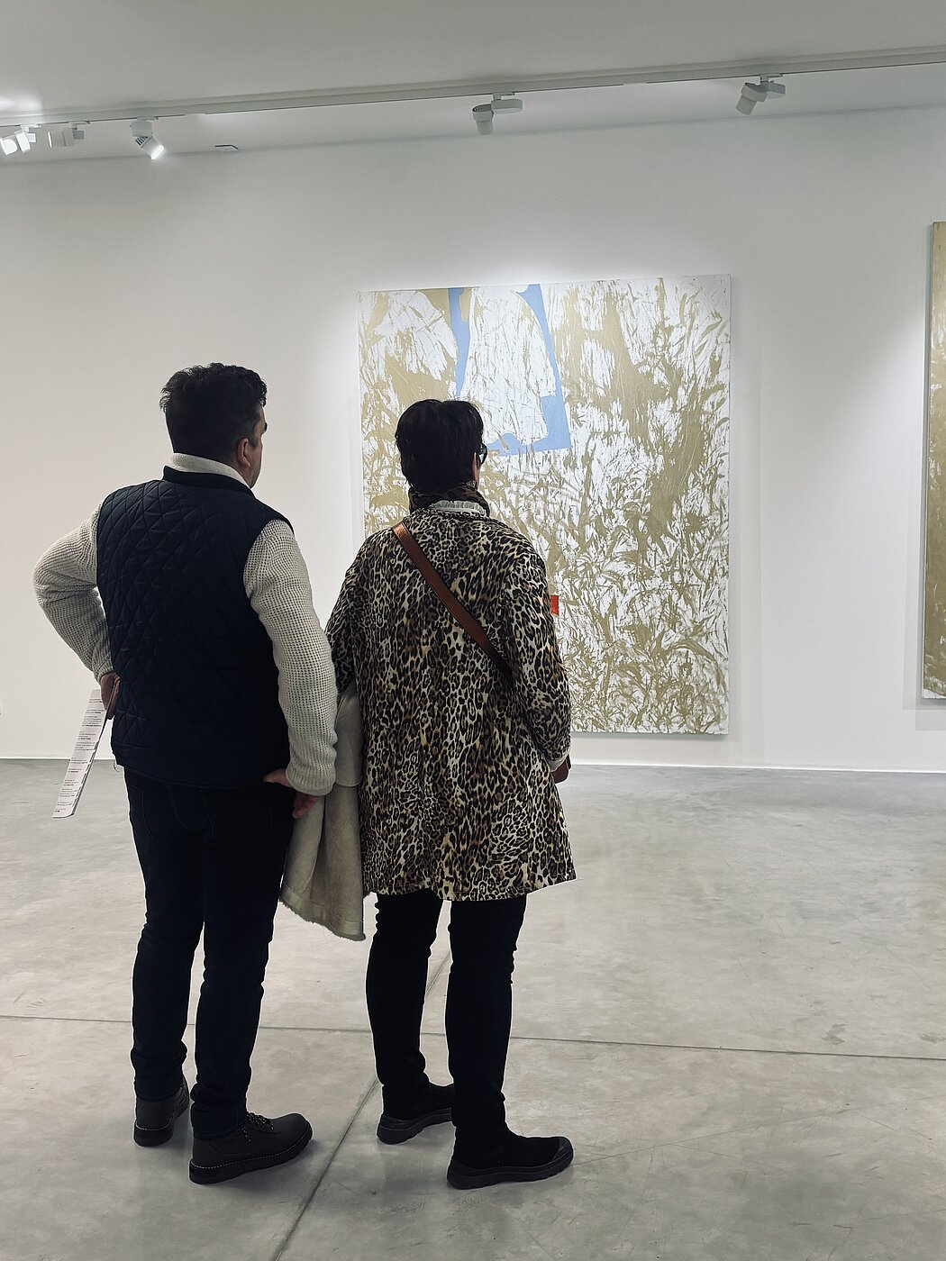     Requête réussie  Gemini a dit  Cette image présente une scène paisible au sein d'une galerie d'art moderne ou d'un musée. Deux personnes, vues de dos, sont en pleine contemplation d'une œuvre d'art de grande dimension.  Voici les éléments clés de la composition : Les Visiteurs      À gauche : Un homme portant un pull en maille claire sous un gilet matelassé bleu marine. Il tient ce qui semble être une brochure ou un programme de l'exposition dans sa main gauche.      À droite : Une femme vêtue d'un manteau à motif léopard (taches brunes et noires sur fond beige). Elle porte un sac en bandoulière et tient un vêtement clair sur son bras gauche.  L'Œuvre d'Art      Il s'agit d'une peinture abstraite verticale fixée au mur blanc.      Le style est gestuel, avec des motifs de couleur or ou ocre qui ressemblent à des branchages ou à des éclaboussures organiques sur un fond blanc.      Une forme géométrique bleue se détache vers le centre supérieur de la toile, apportant un contraste net avec les tons terreux.  Le Cadre      L'espace : Une salle d'exposition minimaliste avec des murs blancs immaculés et un sol en béton poli gris clair.      L'éclairage : Un système de projecteurs sur rails est visible au plafond, dirigeant une lumière diffuse et précise sur les œuvres.      À l'extrême droite, on aperçoit le bord d'une seconde toile, suggérant la continuité de l'exposition.  L'atmosphère générale est celle d'un moment de réflexion tranquille et d'appréciation artistique.