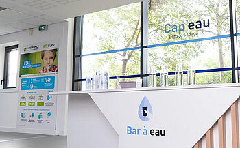 L’image montre un espace intérieur lumineux avec de grandes fenêtres laissant voir des arbres à l’extérieur. À droite, un comptoir blanc porte l’inscription « Bar à eau » avec un logo en forme de goutte d’eau. Sur le comptoir, plusieurs bouteilles et carafes transparentes sont alignées. À gauche, un panneau d’information est fixé au mur, avec des logos, des textes explicatifs et une photographie d’un enfant. Les logos de « Métropole » et « Suez » sont visibles. Le vitrage est floqué du texte « Cap’eau – Espace pédagogique ».