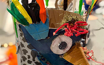 L’image montre un objet fait main à partir de matériaux recyclés ou d'artisanat. Il semble être composé de carton, de papier, de plumes de différentes couleurs (vert, jaune, noir, orange), de boutons, de papier coloré et de fils décoratifs rouges et dorés.  L’objet a une forme cylindrique, avec une structure ressemblant à une tête stylisée. Il y a un œil fait avec du papier blanc et un bouton noir au centre, un bec triangulaire de couleur marron clair, et plusieurs plumes fixées sur le haut. Des motifs noirs et blancs décorent une partie de la surface. Des éléments rouges et dorés sont placés sur le dessus, probablement pour la décoration. On aperçoit des mains et d'autres objets flous en arrière-plan.