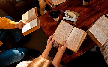 L'image montre une table en bois avec plusieurs livres ouverts. Trois personnes tiennent des livres ouverts, et on peut voir leurs mains et bras, ainsi que des parties de leur corps. Il y a aussi plusieurs tasses blanches et un petit morceau de fromage sur un plat, posé sur la table. Le fond est lumineux, suggérant une lumière naturelle. Les livres sont bien visibles, avec des pages imprimées, et l'atmosphère semble calme et studieuse.