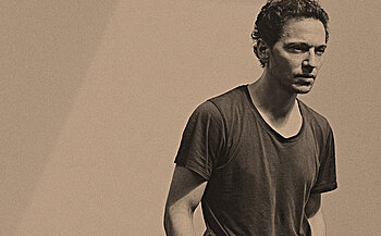 photo de chanteur Raphael L’image montre un homme debout, vêtu d’un t-shirt sombre à col rond. Il a les cheveux courts et bouclés, une barbe naissante, et regarde légèrement vers le bas, avec une expression neutre. L’image est en tons sépia, ce qui lui donne un aspect ancien ou artistique. L’arrière-plan est uni et sans éléments distinctifs.