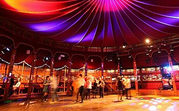 L'image montre un espace intérieur décoré pour une danse. Le plafond est tendu, de couleur rouge avec des lumières violettes et bleues projetées, créant une ambiance festive. Plusieurs personnes dansent au centre de la salle, dont certaines sont floues en raison du mouvement. Des guirlandes lumineuses décorent les murs, et des spots de lumière colorée illuminent le sol en bois. Le lieu semble être un chapiteau ou une salle de danse avec une atmosphère animée et festive.