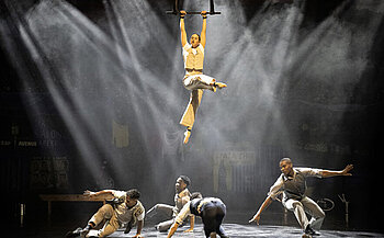 L’image montre une scène de spectacle avec six personnes en action. Une personne est suspendue en l’air, accrochée à une barre tenue par les mains, les jambes croisées, éclairée par un faisceau lumineux. En dessous, cinq autres personnes sont au sol dans différentes postures dynamiques. Elles portent des tenues similaires de couleur claire, avec chemises et pantalons. L’arrière-plan est sombre avec des effets de lumière et de fumée, et des éléments de décor visibles tels que des panneaux et du linge suspendu.