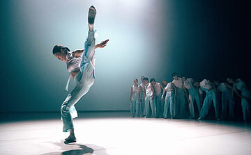 L’image montre une personne en premier plan effectuant un grand mouvement de danse, avec une jambe levée très haut, presque verticale, et les bras étendus. Elle porte un costume clair, dans des tons bleus ou blancs. En arrière-plan, un groupe de personnes se tient aligné, observant la scène ou penché dans une posture similaire. Elles portent également des tenues claires. La scène est baignée d’un éclairage bleuté et tamisé, créant une ambiance douce et diffuse. Le sol est clair et l’espace semble vaste.
