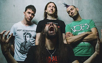 Photo Ultra Vomit  - L’image montre quatre personnes posant devant un mur gris. Trois d’entre elles sont debout à l’arrière-plan, les bras croisés ou le long du corps, tandis qu'une personne est au premier plan, criant ou ouvrant grand la bouche, les bras levés. Les individus portent des t-shirts à motifs ou logos visibles, et l’un d’eux a une crête colorée. L’expression faciale de la personne au premier plan est très expressive, contrastant avec les visages plus neutres ou légèrement souriants des autres.