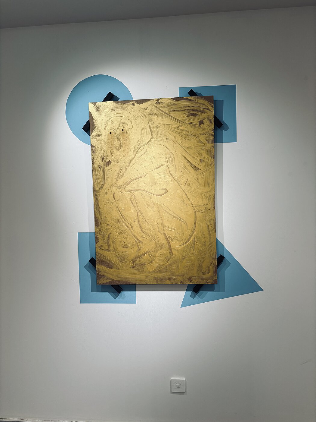 Cette œuvre d'art contemporaine présente un mélange fascinant de textures organiques et de formes géométriques rigides. Voici une description détaillée de la pièce : L'élément central : Le panneau doré  Au cœur de l'installation se trouve un panneau rectangulaire vertical recouvert d'une peinture dorée ou ocre métallique.      La technique : La peinture semble avoir été appliquée de manière gestuelle et texturée, avec des coups de pinceau larges et tourbillonnants qui créent du relief.      Le sujet : Dans les textures de la peinture, on devine une forme anthropomorphe ou animale (évoquant peut-être un singe ou un personnage accroupi) avec deux points sombres en guise d'yeux en haut à gauche. L'aspect est volontairement brut et primitif.  La mise en scène géométrique  Le panneau ne repose pas directement sur le mur, mais s'intègre dans une composition graphique plus large :      Formes bleues : Derrière le panneau, des formes géométriques d'un bleu plat et mat (un cercle, un carré et un triangle) sont peintes ou fixées au mur. Elles créent un contraste frappant avec l'aspect "sale" et texturé du panneau doré.      Fixations : Le panneau est maintenu par huit tasseaux noirs disposés par paires à chaque coin, rappelant des pinces ou des agrafes industrielles. Cela donne l'impression que l'œuvre est "clouée" ou exposée de force sur ses supports géométriques.  L'ambiance générale  L'ensemble joue sur les contrastes :      Brut vs Lisse : Le panneau texturé et doré s'oppose à la planéité parfaite des formes bleues.      Ordre vs Chaos : La rigueur des mathématiques (cercle, carré, triangle) tente de contenir une figure organique et sauvage.