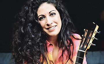 L’image montre une personne aux cheveux longs, noirs et bouclés, vêtue d’une chemise colorée dans les tons rose et orange. Elle est assise et sourit en regardant vers l’objectif. Elle tient une guitare acoustique, dont le manche et une partie de la tête sont visibles à droite de l’image. L’arrière-plan est sombre, mettant en valeur le visage et les cheveux de la personne.