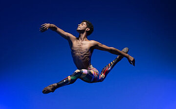 Corps de danseur tendu dans une position classique de grand jeté, une figure emblématique du ballet, où une jambe est projetée vers l'avant et l'autre vers l'arrière dans un élan parfaitement maîtrisé.  Le danseur, torse nu, révèle une musculature fine et sculptée -  Il porte un collant aux couleurs vives et géométriques, qui contraste avec le fond bleu profond et l’éclairage dramatique qui souligne les contours de son corps.  Son regard est tourné vers le haut L’ensemble dégage une sensation de liberté, de puissance maîtrisée et d’élégance aérienne.