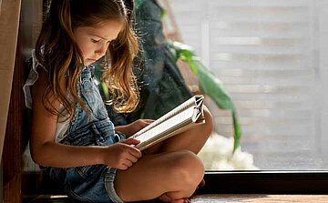 fillette assises avec des couettes lisant un livre avec une plante verte en fond 