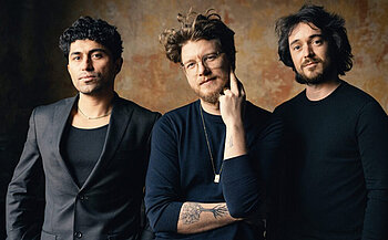 Les musiciens Marck Priore, Juan Villarroel, et Élie Martin-Charrière - L’image montre trois personnes debout, posant face à l’objectif. La personne à gauche porte un costume noir avec un t-shirt foncé. Celle au centre porte un haut à manches longues foncé, des lunettes, un collier doré, et a des tatouages visibles sur l'avant-bras gauche. Elle touche son visage avec sa main droite. La personne à droite porte un haut noir à manches longues. L’arrière-plan est texturé avec des teintes chaudes.