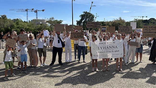 Cette photo montre un rassemblement de personnes, majoritairement vêtues de blanc, participant à une manifestation pacifique en extérieur sous un ciel bleu et dégagé. Au premier plan, trois jeunes filles tiennent une grande banderole blanche sur laquelle est inscrit : « MARCHE BLANCHE EN HOMMAGE À LYHANNA