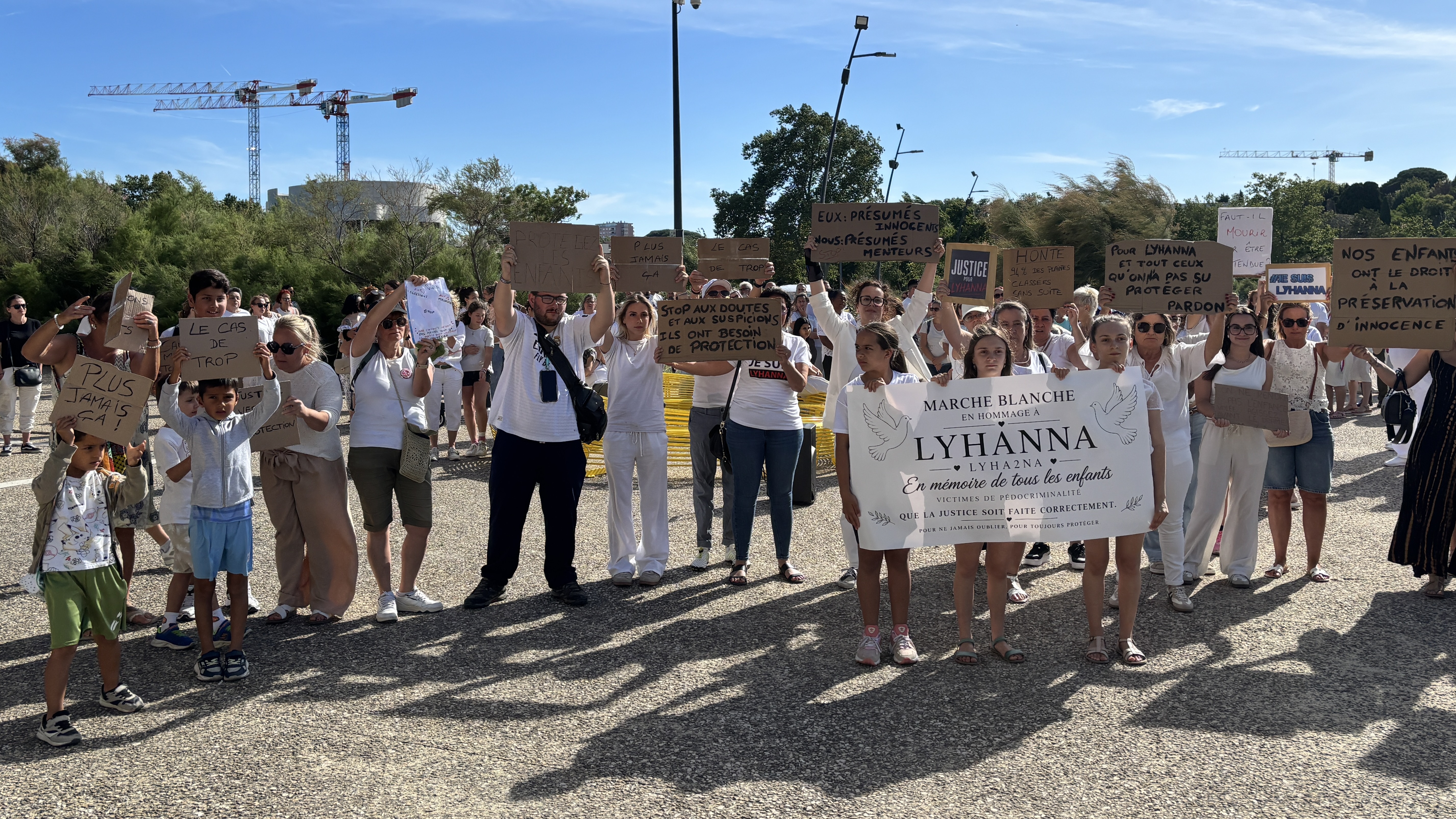 Cette photo montre un rassemblement de personnes, majoritairement vêtues de blanc, participant à une manifestation pacifique en extérieur sous un ciel bleu et dégagé. Au premier plan, trois jeunes filles tiennent une grande banderole blanche sur laquelle est inscrit : « MARCHE BLANCHE EN HOMMAGE À LYHANNA 