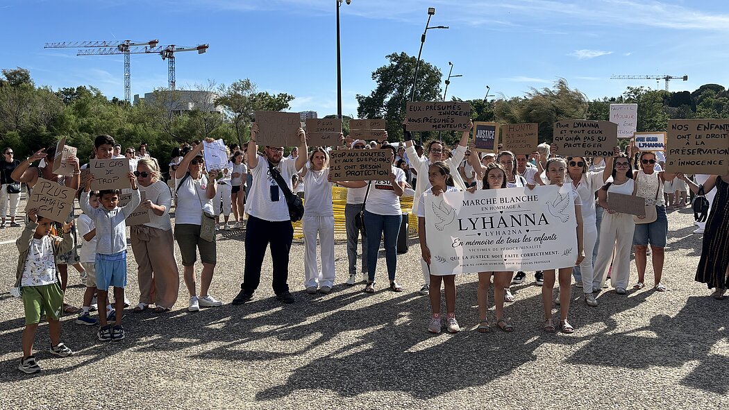 Cette photo montre un rassemblement de personnes, majoritairement vêtues de blanc, participant à une manifestation pacifique en extérieur sous un ciel bleu et dégagé. Au premier plan, trois jeunes filles tiennent une grande banderole blanche sur laquelle est inscrit : « MARCHE BLANCHE EN HOMMAGE À LYHANNA 