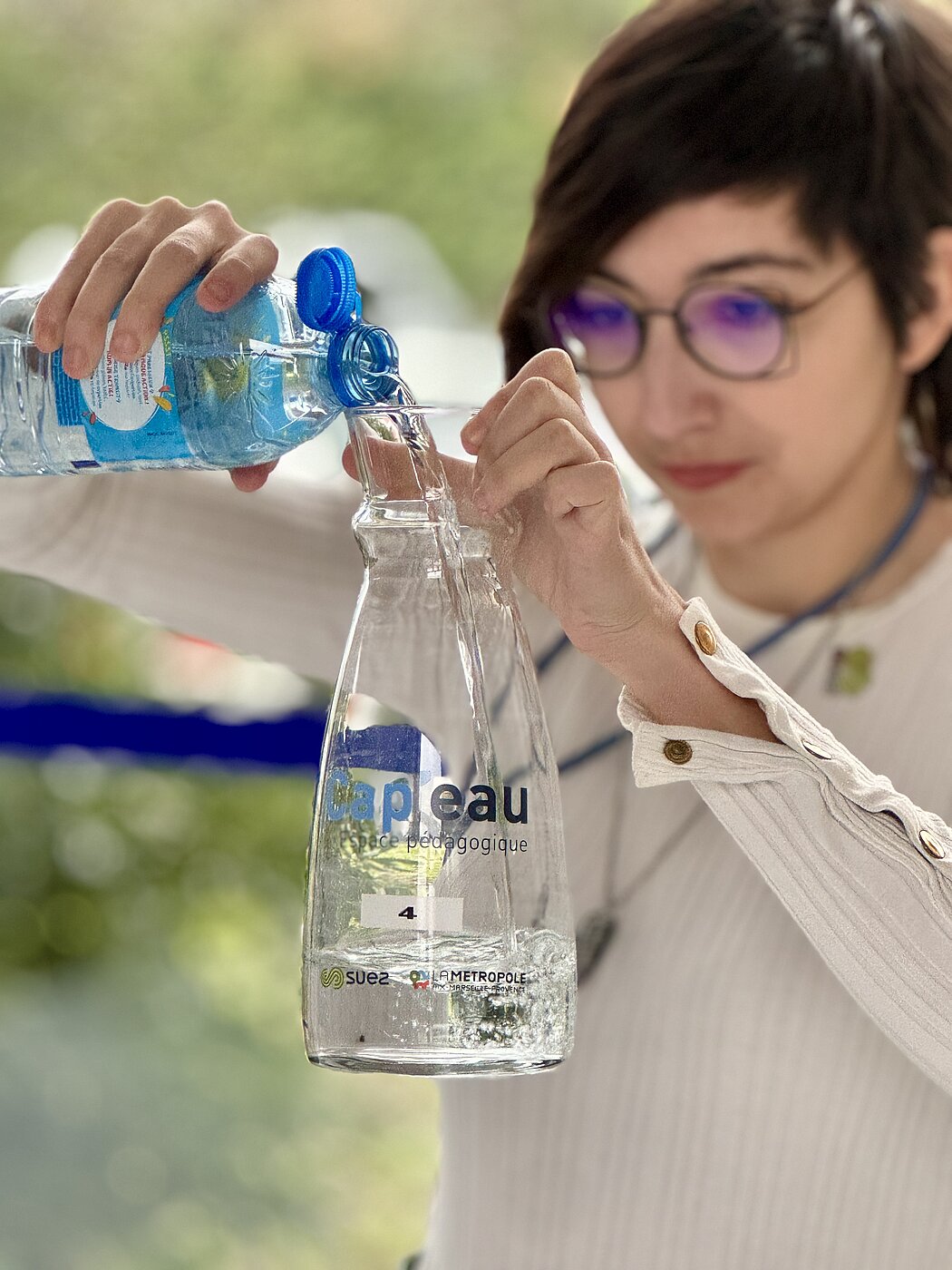 Cette photo montre une femme versant de l'eau d'une bouteille en plastique bleu dans une carafe en verre transparente.  Voici les éléments clés de l'image :  Action principale : Une personne, portant des lunettes et un haut blanc à manches longues, remplit une carafe.  La carafe : Elle porte l'inscription "Cap l'eau espace pédagogique", accompagnée des logos de Suez et de La Métropole Aix-Marseille-Provence. Le chiffre "4" est également visible sur une étiquette.  Contexte : Le flou en arrière-plan suggère un environnement extérieur ou très lumineux, mettant l'accent sur le geste et le message lié à la gestion de l'eau ou à la sensibilisation pédagogique.