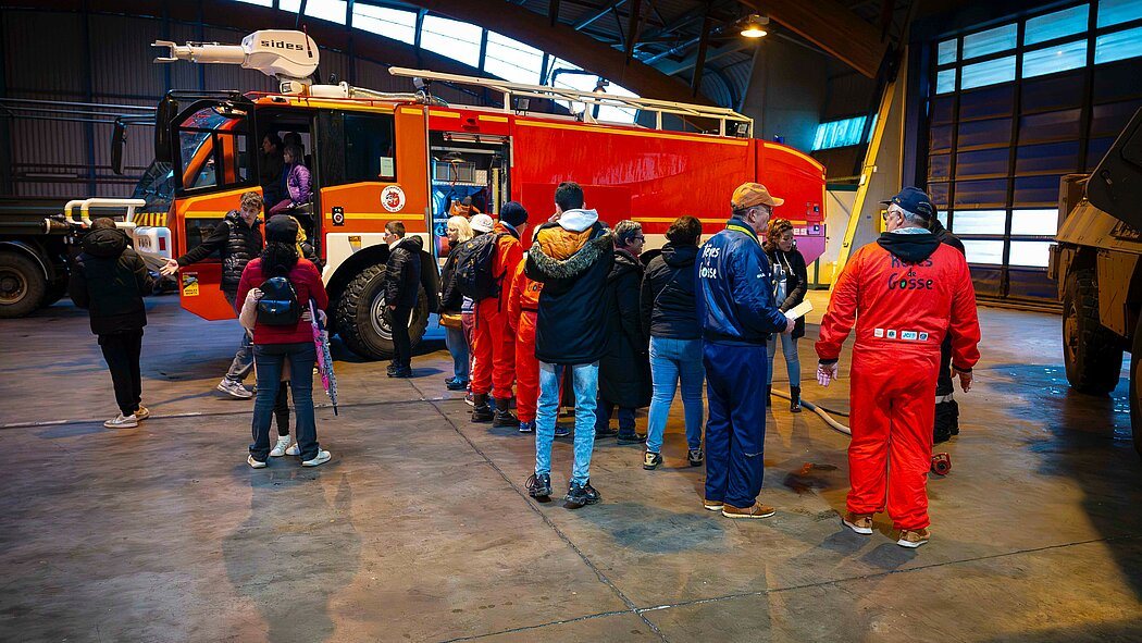 Cette image montre une scène animée à l'intérieur d'un grand hangar de la  base aérienne centrée autour d'un imposant camion de pompiers rouge. Le camion est équipé d'un canon à eau sur le toit. On peut lire l'inscription "Pompiers de l'Air" sur le côté de la cabine. Les portes de la cabine et des compartiments latéraux sont ouvertes, permettant aux visiteurs d'en découvrir l’intérieur. Un groupe de personnes, composé d'adultes et d'enfants, est rassemblé autour du véhicule. Plusieurs personnes portent des vestes bleues ou des combinaisons rouges avec l'inscription "Rêves de Gosse". Il s'agit d'une association française qui organise des événements aéronautiques pour favoriser l'inclusion entre enfants "extraordinaires" (touchés par le handicap ou la maladie) et enfants "ordinaires ». On voit des enfants monter dans la cabine du camion ou attendre leur tour, tandis que des adultes les encadrent ou discutent entre eux. L'atmosphère semble être celle d'une journée de découverte et de partage.