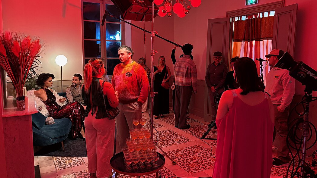 Cette image nous plonge au cœur d'un plateau de tournage sophistiqué, installé dans une pièce élégante au style classique. L'atmosphère est dominée par une lumière rouge vive et artistique qui crée un contraste saisissant avec les éléments de décor plus sombres.