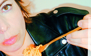 Photo de la chanteuse Mademoiselle K L’image montre une personne partiellement cadrée, principalement sur le côté gauche de l’image. Elle porte une veste en cuir noir avec des boutons métalliques. Elle tient une fourchette contenant des spaghettis. Le fond est clair et uni. La personne regarde vers l’objectif avec une expression neutre.